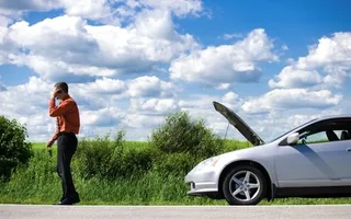 A man on a road talking on his phone while a white car with an open hood is stopped nearby